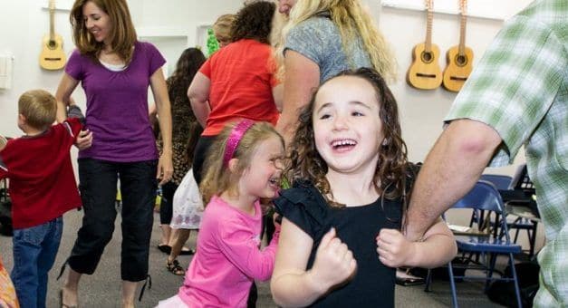 Children laughing and dancing with their parents in a Children's Music Academy class, with guitars hanging on the wall.