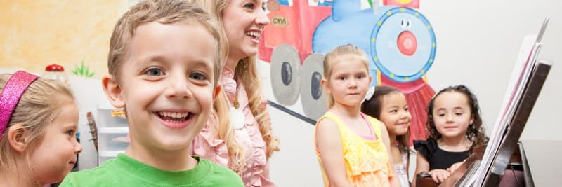 A smiling boy in front of a class of young children and their teacher gathered at the piano.