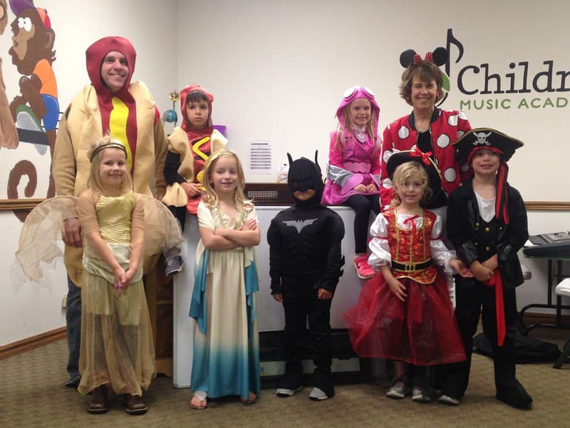 Children in costumes with their teacher and parents in front of the Children's Music Academy sign in the classroom.