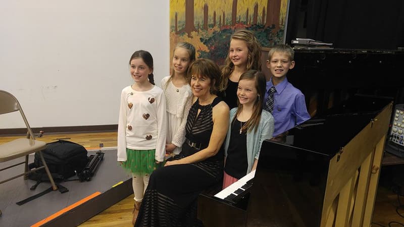 A teacher seated at the piano with a group of older CMA students around her.
