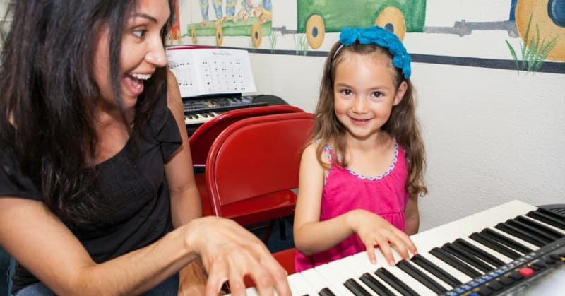 A smiling teacher and a young girl in a pink dress playing keyboards together in a Children's Music Academy classroom.